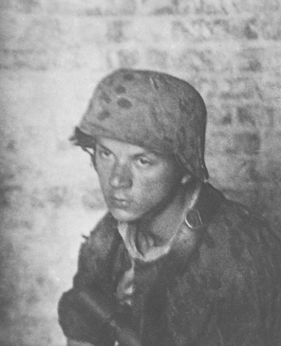 Młody mężczyzna o poważnej twarzy, ubrany w mundur i hełm, z bronią przewieszoną przez ramię