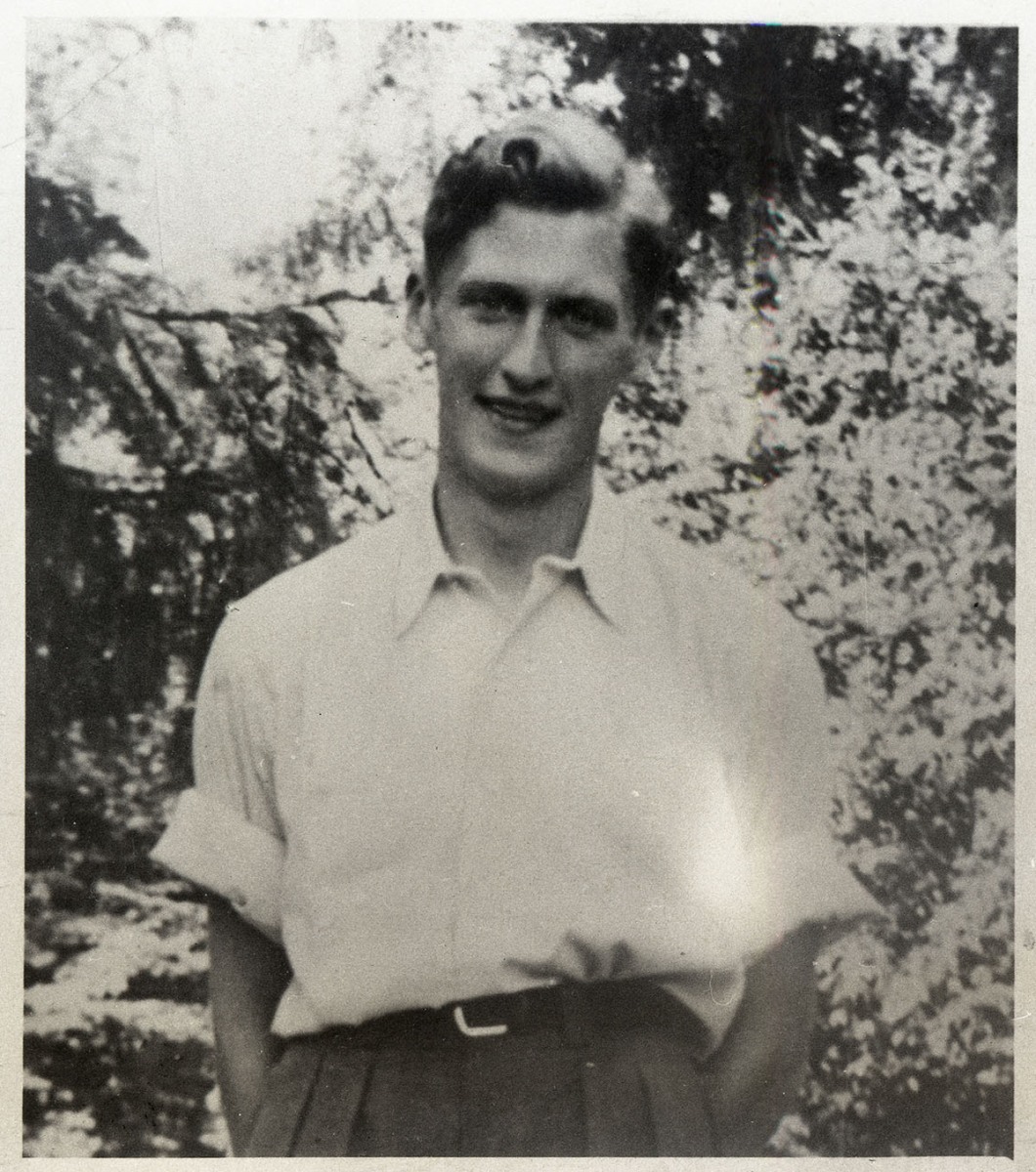 Uśmiechnięty młody mężczyzna, stojący na tle drzew z rękami w kieszeniach, ubrany w luźną koszulę