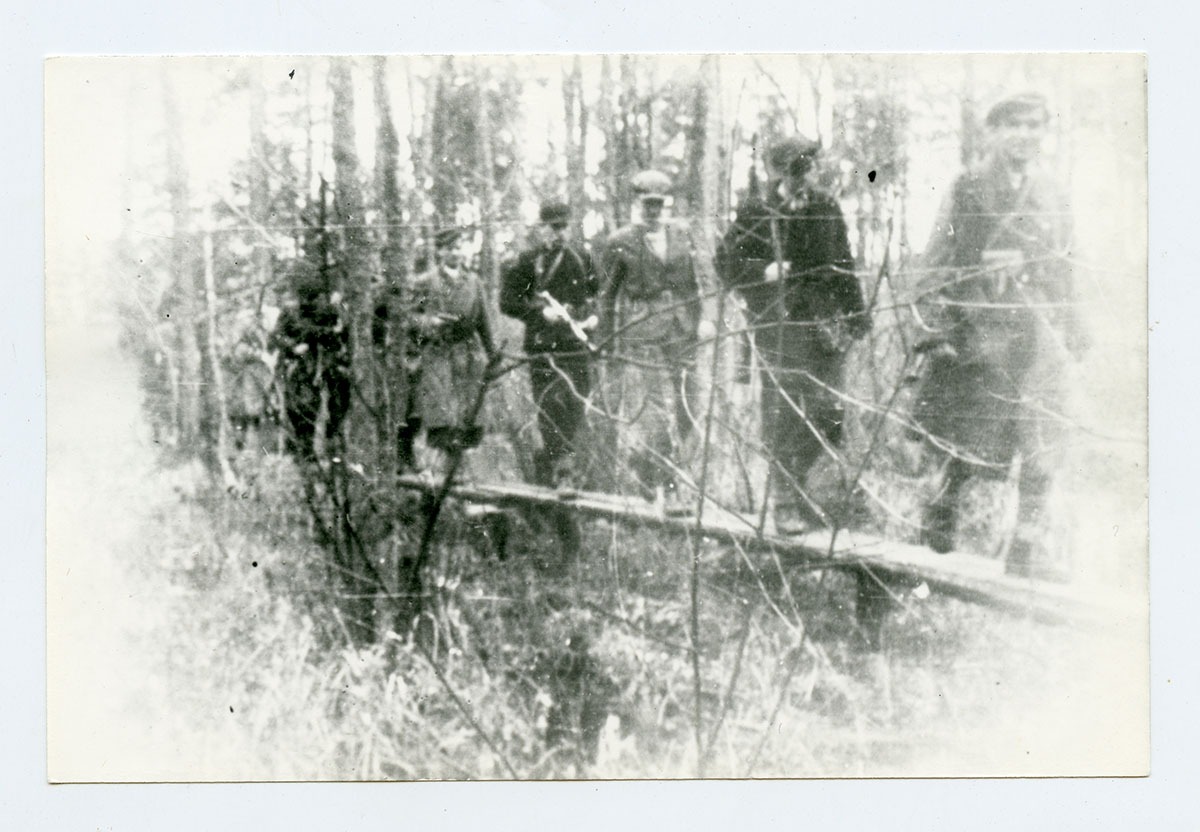 grupa uzbrojonych mężczyzn, ubranych po cywilnemu, przechodzących wąską kładką w lesie