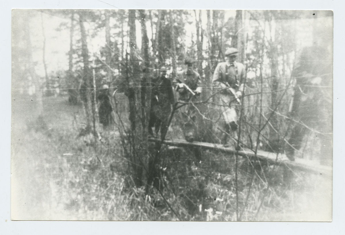 grupa uzbrojonych mężczyzn, ubranych po cywilnemu, przechodzących wąską kładką w lesie