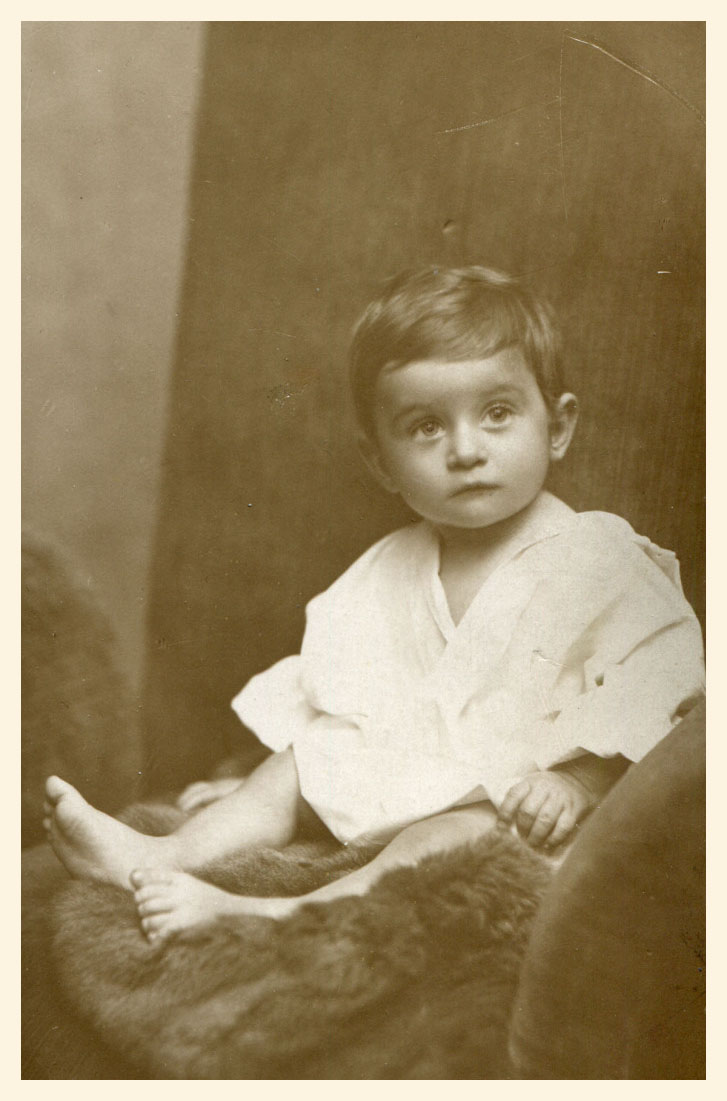 Chłopczyk około roku, siedzący w fotelu na kawałku futra w koszulce, spod której widać gołe nóżki