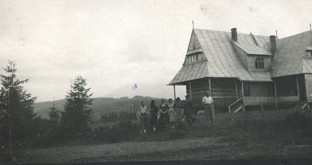 Parterowy dom góralski z górami w tle; przed nim 7 młodych osób – trzej mężczyźni i 4 kobiety