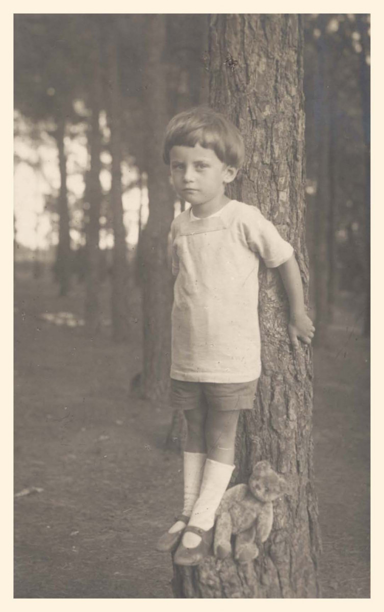 Chłopczyk około 4 lat stoi oparty o pień drzewa w lesie; u jego stóp siedzi pluszowy miś