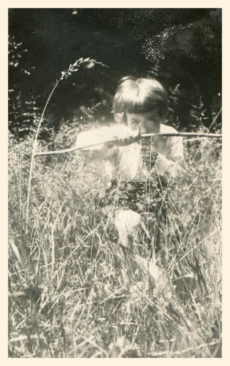 Chłopiec około 7 lat, klęczący na jedno kolano w wysokiej trawie i celujący z łuku-zabawki na wprost