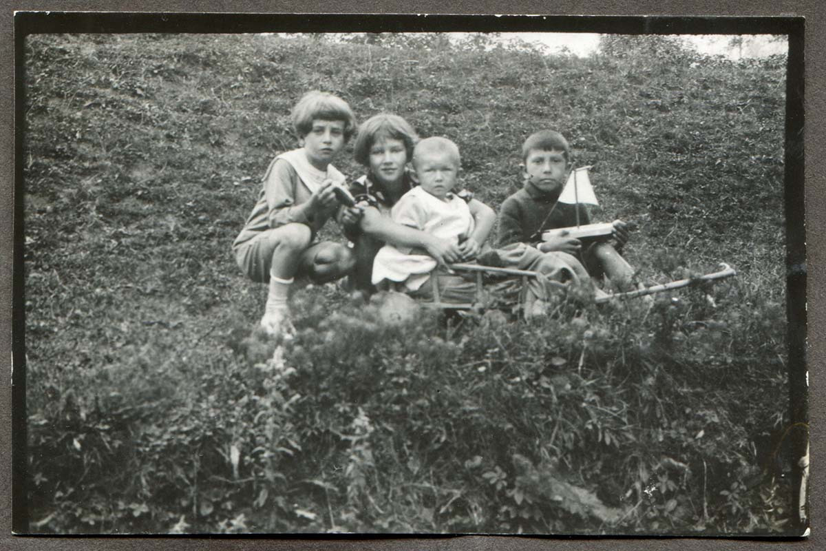Na trawie dwaj chłopcy i dziewczynka około 7 lat, przed nią małe dziecko w drewnianym wózku