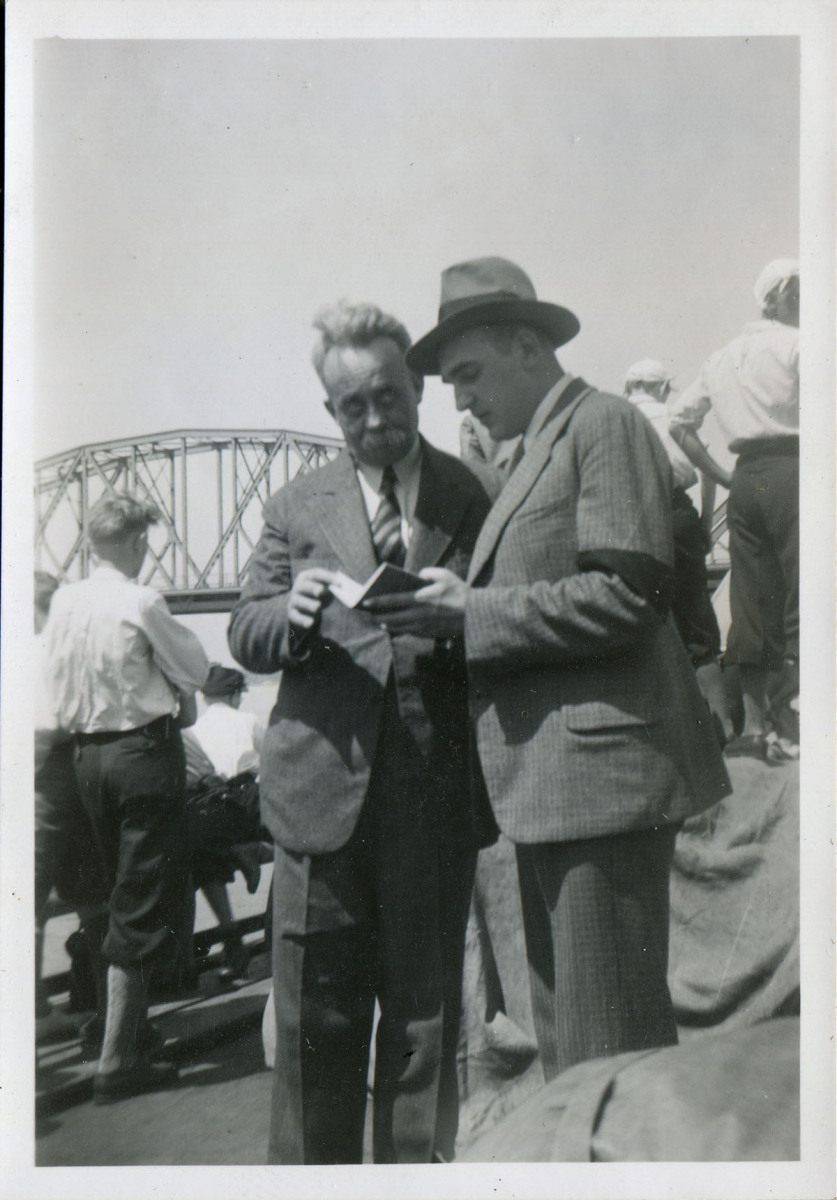 Dwaj mężczyźni, zaglądający razem do notesu. Za nimi tyłem chłopcy, patrzący na most w tle