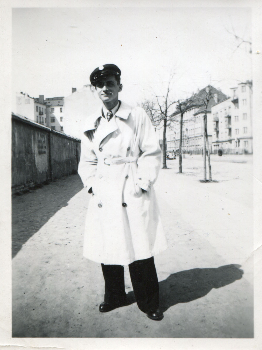 Młodzieniec w kaszkiecie z emblematem szkoły, stojący na ulicy z rękami w kieszeniach płaszcza