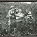 Na trawie dwaj chłopcy i dziewczynka około 7 lat, przed nią małe dziecko w drewnianym wózku