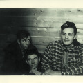 Trzej chłopcy około 17 lat, siedzący pod ścianą z desek, dwaj widoczni do pasa, środkowy do ramion